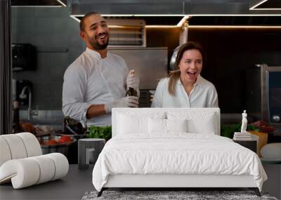 Handsome young African chef is cooking together with his Caucasian girlfriend in the kitchen using red wine as an ingredient. Wall mural