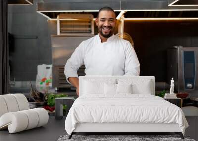 Handsome young African chef standing in professional kitchen in restaurant preparing a meal of meat and cheese vegetables. Wall mural