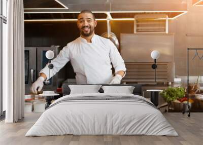 Handsome young African chef standing in professional kitchen in restaurant preparing a meal of meat and cheese vegetables. Wall mural