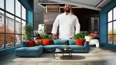 Handsome young African chef standing in professional kitchen in restaurant preparing a meal of meat and cheese vegetables. Wall mural