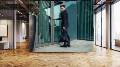 handsome young businessman with a beard and in a business suit standing on the street against the background of the office building next to a comfortable stylish leather bag. Wall mural