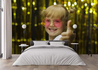 Child show thumbs up, smiling, looking at camera. Girl posing on background with foil golden curtain Wall mural