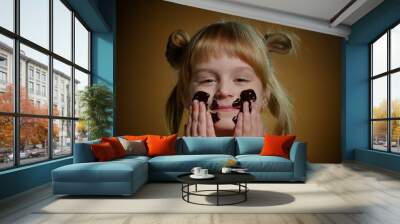 Close-up portrait of happy teen child kid smears her face with melted dark chocolate and starts eating chocolate bar. Satisfied young blond girl with two ponytails smiling on dark brown background Wall mural