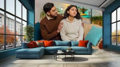 Compassion. Caring young man comforting upset Indian girlfriend sitting on sofa in room at home. Husband and wife family marriage couple together on couch in apartment. Man supporting depressed woman. Wall mural