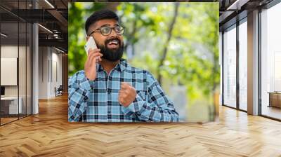 Happy Indian bearded man having remote conversation communicate speaking by smartphone outdoors. Hispanic guy talking on phone unexpected good news gossip on city street. Town lifestyles. Horizontal Wall mural