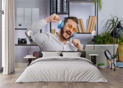 Happy relaxed overjoyed middle eastern businessman working on laptop computer at office wearing headphones listening favorite energetic disco music and dancing. Freelancer man relaxing, taking a break Wall mural