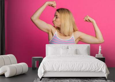 I am strong and independent. Caucasian young woman showing biceps and looking confident, feeling power strength to fight for rights, energy to gain success win. Blonde girl isolated on pink background Wall mural