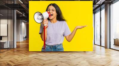 Indian young woman talking with megaphone, proclaiming news, loudly announcing advertisement discounts sale, using loudspeaker to shout speech. Happy Arabian girl isolated on studio yellow background Wall mural