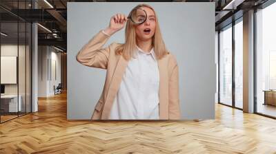 Investigator researcher scientist office woman holding magnifying glass near face, looking into camera with big zoomed funny eyes, searching analysing. Business girl isolated on gray studio background Wall mural