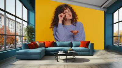 Need some more, please give me. Curly haired woman showing a little bit gesture with sceptic smile, showing minimum sign, measuring small size. Young girl isolated alone on yellow studio background Wall mural