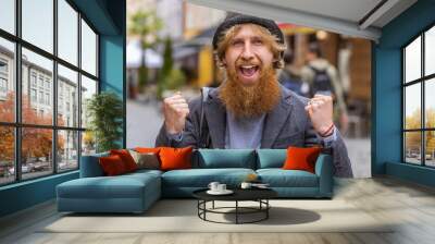 Portrait of young man shouting, celebrating success, winning, goal achievement, good news, lottery jackpot luck, victory outdoors. Redhead bearded guy walking in urban city street. Town lifestyles Wall mural