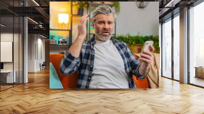 Shocked sad mature man holding smartphone reading negative message feels annoyed sitting on sofa in living room. Caucasian senior elderly old guy having gadget trouble in apartment. App crash concept. Wall mural