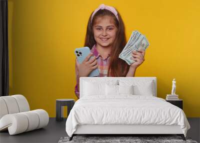 Smiling happy young child girl kid looking smartphone display sincerely rejoicing win, receiving money dollar cash banknotes, success lottery jackpot luck. Little toddler children on yellow background Wall mural