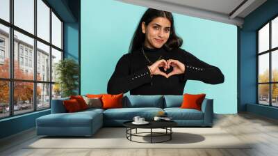 Woman in love. Smiling arabian indian woman 20 years old makes heart gesture demonstrates love sign expresses good feelings and sympathy. Pretty hindu girl isolated alone on blue studio background Wall mural