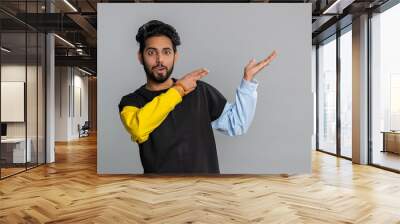 Young indian man showing thumbs up and pointing empty place, advertising area for commercial text, copy space for goods promotion. Bearded hindu guy isolated alone on gray studio background, indoors Wall mural