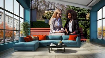 Two businesswomen sitting on the bench and looking at the tablet screen. One women is black. Wall mural