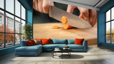 A man cuts fresh carrots with a large knife on a wooden table preparing to eat Wall mural