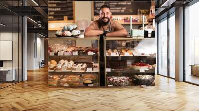 Cheerful indian male baker welcoming you at his bakery store, bearded male baker smiling to the camera proudly leaning on the showcase at his cafe Wall mural