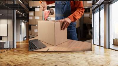 Worker man In Retail Warehouse working packing parcel box with tape dispenser for shipment. Delivery and distribution center full of shelves with goods. Close-up Wall mural