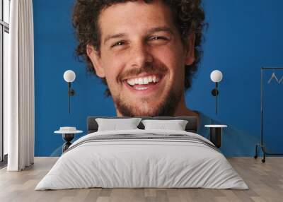 Close up, guy with curly hair dressed in blue t-shirt looks at the camera and laughs isolated on blue background in the studio Wall mural