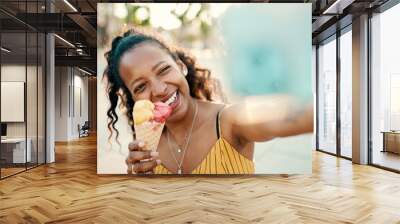 Closeup of smiling young woman with long curly hair with ice cream in her hands making video call on a mobile phone on an urban city background. Close-up of happy girl using smartphone to communicate Wall mural