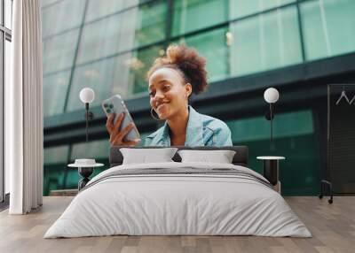 Cute African girl with ponytail, wearing denim jacket, in crop top with national pattern, sitting on bench and using smartphone on modern buildings background. Wall mural