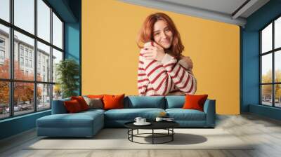 Cute red-haired woman dressed in sweater hugs herself looking at camera on yellow background. Beautiful girl poses in studio with blank space on yellow wall for your promotion Wall mural