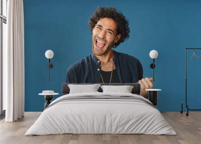 Happy attractive man with curly hair, wearing blue shirt, looking at camera surprised wow raises his hands celebrating victory isolated on blue background in studio Wall mural