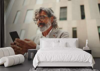 Mature businessman with beard in eyeglasses wearing gray jacket is using cell phone. Middle aged manager scrolling information on his smartphone while sitting outside the office Wall mural