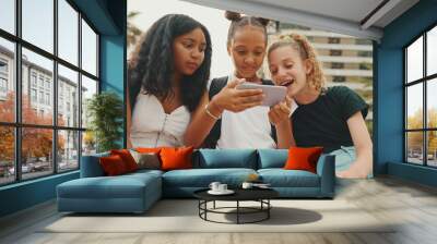 Three girls friends pre-teenage are sitting on the waterfront using mobile phone, watching videos, photos. Three teenagers on the outdoors in urban cityscape background Wall mural