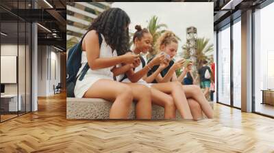 Three girls friends pre-teenage are sitting on the waterfront using mobile phone. Three teenagers playing games on a smartphone on the outdoors in urban cityscape background Wall mural