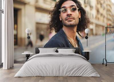 Young italian guy with long curly hair and stubble walks smiling down the street. Stylish man with sunglasses and lot of chains smiling and looking around Wall mural