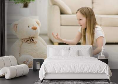 Photo of little blond girl talking to her white teddy bear. Wall mural