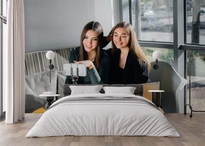 Two young beautiful girls are sitting in a cafe, recording video blogs and communicating on social networks. Wall mural