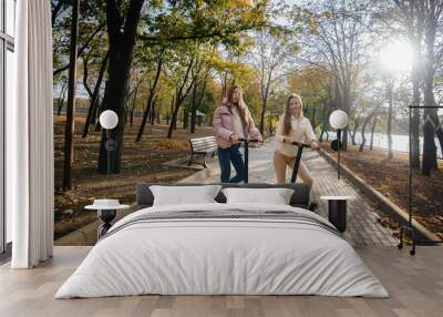 Two young beautiful girls ride electric scooters in the Park on a warm autumn day Wall mural