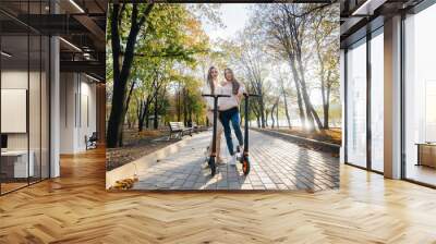 Two young beautiful girls ride electric scooters in the Park on a warm autumn day Wall mural