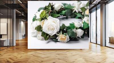 A flower crown or wreath made of white roses and green leaves, arranged in a circular pattern, on a plain white surface, shot from a slightly elevated angle. Wall mural