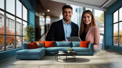 A man and a woman are smiling and holding a tablet. The man is wearing a suit and the woman is wearing a pink jacket. They seem to be happy and enjoying their time together Wall mural