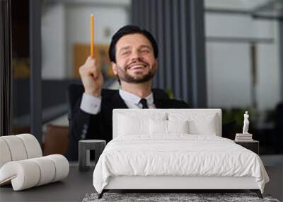 A man in a suit is holding a pencil and smiling. He is sitting at a desk with a notebook and a book. Concept of productivity and focus, as the man is likely working on a project or studying Wall mural