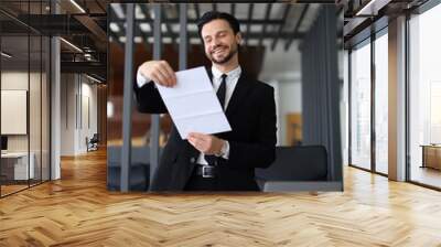 A man in a suit is holding a piece of paper and smiling. He is standing in front of a desk with a laptop and a chair. Concept of professionalism and confidence Wall mural