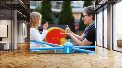 A woman and a child communicate with sign language. They are sitting on a blue playground in an outdoor setting. They are mother and daughter or teacher and student. Wall mural