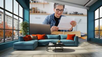 Happy mature woman watching mail bank notice, sitting at kitchen table at home. Wall mural