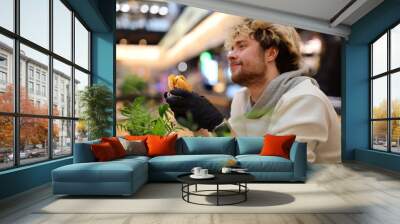 Hungry man holding a cheeseburger in his hand. He is sitting at a counter in a cafe with plants in the background. Wall mural