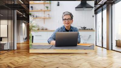 Mature woman working on a laptop in a home interior on the background of the kitchen. Wall mural