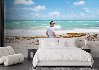 Young happy man smiling standing on sand sandy beach in Miami, Florida with turquoise blue green ocean sea waves background of flying pelican bird Wall mural