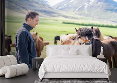 Young happy man standing, watching many Icelandic horses in outdoor stable paddock, Iceland in countryside rural farm, mountains, making funny faces, showing teeth, open mouth Wall mural