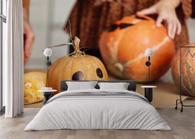 Close up of young woman carving Jack O Lattern from ripe orange pumpkin with knife on her wooden kitchen table. Female preparing all hallows eve Halloween party decorations. Background, copy space. Wall mural