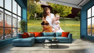 Family day, mother's day. Beautiful smiling young mom and baby daughter cuddling happy domestic dog on the backyard lawn.Mother with child girl are having fun with pet outdoors on summer holiday. Wall mural