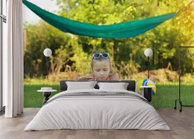 kid girl is eating piece of pizza and playing on laptop lying on grass on summer sunny day. the child has lunch after active games. hammock and ball on the background. Wall mural