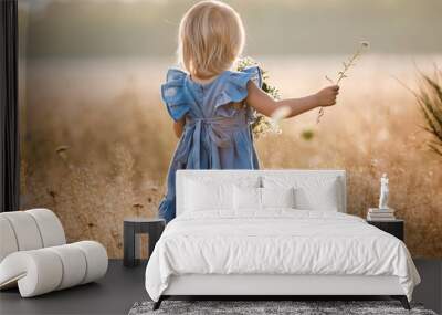 little girl is wearing in a blue dress in a field on summer sunny day. Portrait of adorable little child outdoors. happy holiday childhood. back view. Wall mural